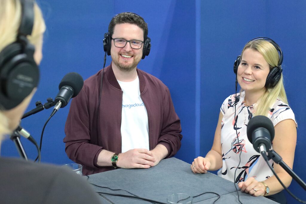 Zwei Personen im Podcast-Studio zwischen mehreren Mikrofonen vor blauem Hintergrund sprechen zum Thema "Wie gelingt Elektromobilität im Alltag?".